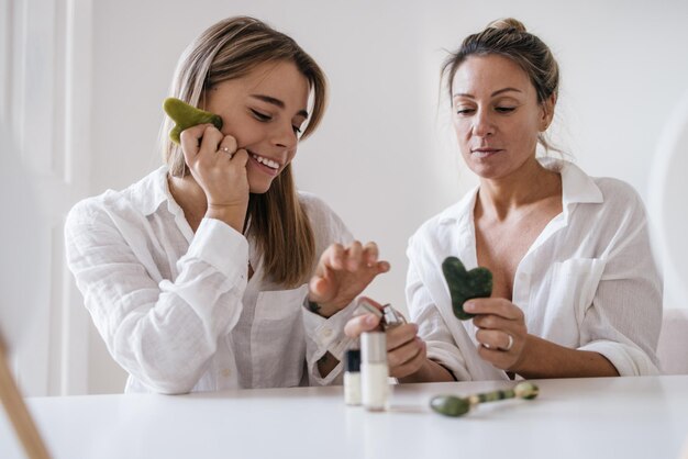 Zwei kaukasische blonde Frauen unterschiedlichen Alters probieren kosmetische Produkte, während sie auf weißem Hintergrund sitzen. Hautpflege- und Feuchtigkeitskonzept