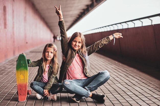 Zwei kleine fröhliche Schwestern sitzen auf dem Skateboard im Tunnel.