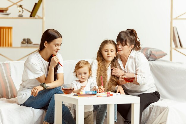 Zwei kleine Mädchen, ihre attraktive junge Mutter und ihre charmante Großmutter, sitzen auf dem Sofa und verbringen Zeit miteinander zu Hause. Generation von Frauen. Internationaler Frauentag. Schönen Muttertag.