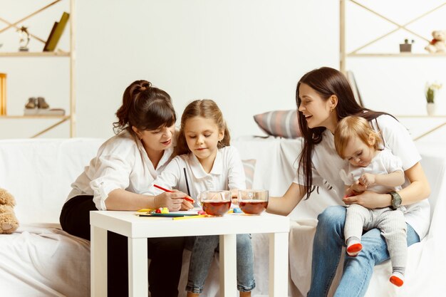 Zwei kleine Mädchen, ihre attraktive junge Mutter und ihre charmante Großmutter, sitzen auf dem Sofa und verbringen Zeit miteinander zu Hause. Generation von Frauen. Internationaler Frauentag. Schönen Muttertag.