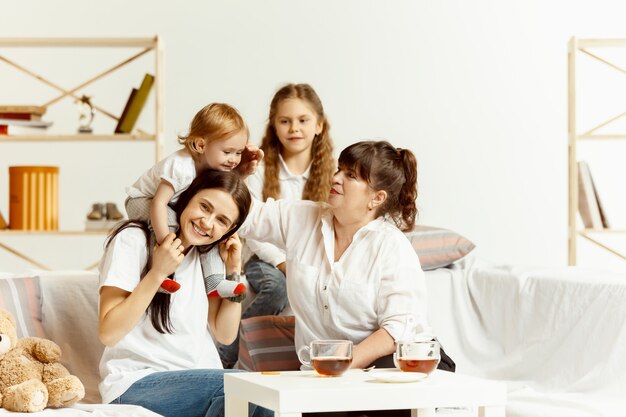 Zwei kleine Mädchen, ihre attraktive junge Mutter und ihre charmante Großmutter, sitzen auf dem Sofa und verbringen Zeit miteinander zu Hause. Generation von Frauen. Internationaler Frauentag. Schönen Muttertag.