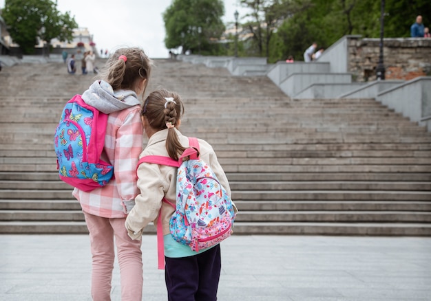 Zwei kleine Mädchen mit Rucksäcken auf dem Rücken gehen Hand in Hand zur Schule. Kindheitsfreundschaft