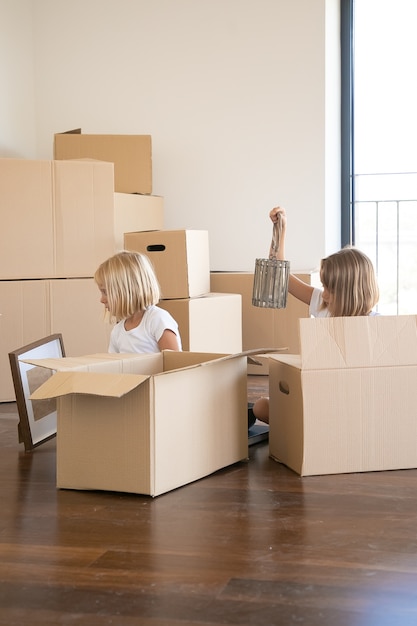 Zwei kleine Mädchen packen Dinge in der neuen Wohnung aus, sitzen auf dem Boden und nehmen Gegenstände aus offenen Comicboxen