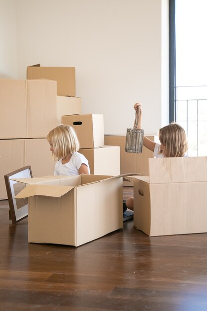 Zwei kleine Mädchen packen Dinge in der neuen Wohnung aus, sitzen auf dem Boden und nehmen Gegenstände aus offenen Comicboxen