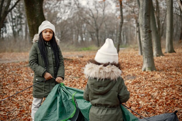 Zwei kleine schwarze Mädchen beim Zeltcamping im Wald Zwei kleine Schwestern bauen im Herbstwald ein Zelt auf Schwarze Mädchen in Khaki-Mänteln und beigen Hüten
