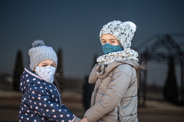 Zwei kleine Schwestern in wiederverwendbaren Masken und Hüten während der Quarantänezeit auf dunklem Hintergrund.