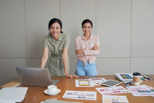 Zwei kreative asiatische Frauen, die im Büro, mit Laptop, Dokumenten und Bildern auf Tabelle aufwerfen