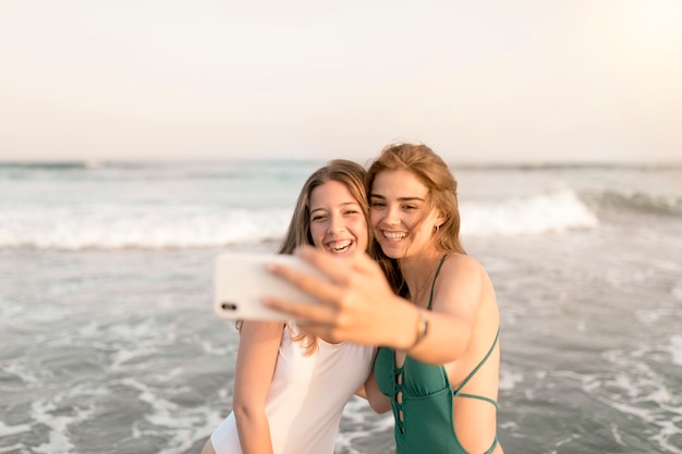 Zwei lächelnde Mädchen, die Selbstporträt vom Handy nahe der Küste nehmen