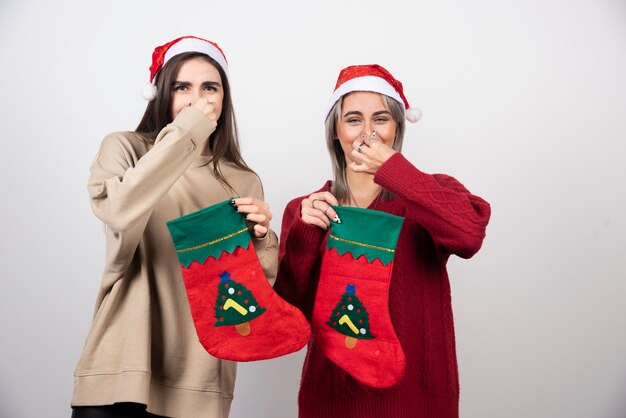 Zwei lächelnde Mädchen in Weihnachtsmütze mit Weihnachtssocken, die ihre Nasen vor Ekel schließen.