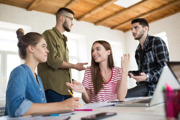 Zwei lächelnde Mädchen unterhalten sich fröhlich, während sie Zeit im modernen Büro mit Kollegen im Hintergrund verbringen Gruppe kreativer Menschen, die zusammenarbeiten