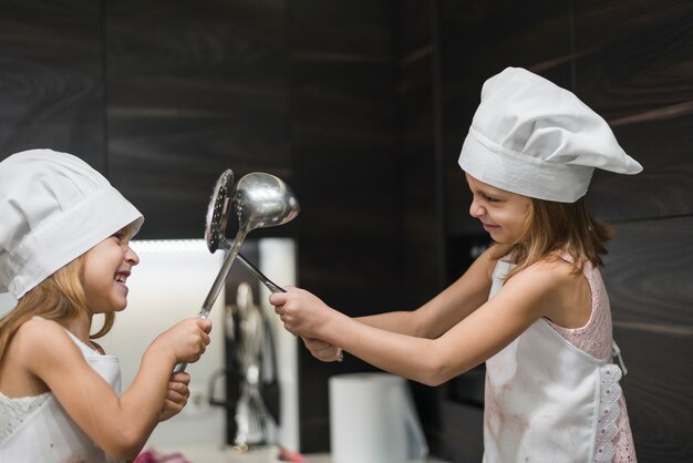Zwei lächelnde nette Schwestern in den Hüten des Chefs kämpfen mit Küchengerät