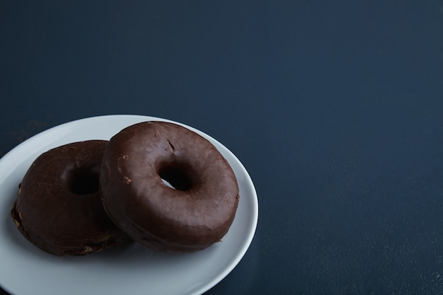 Zwei leckere frisch gebackene Donuts, glasiert mit Schokolade auf weißem Keramikkleinteller lokalisiert in der Ecke des rustikalen alten blauen Holztisches