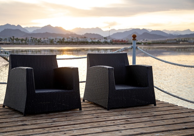 Zwei leere Stühle auf einem hölzernen Pier mit Blick auf die Berge im Sonnenuntergangslicht.