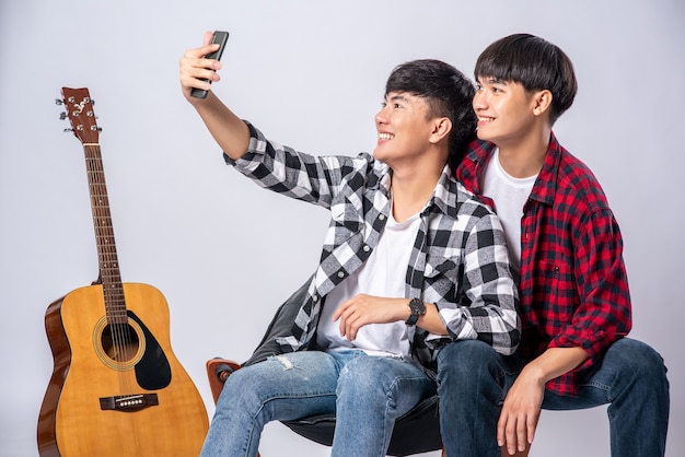 Zwei liebevolle junge Männer sitzen auf einem Stuhl und machen ein Selfie von einem Smartphone.