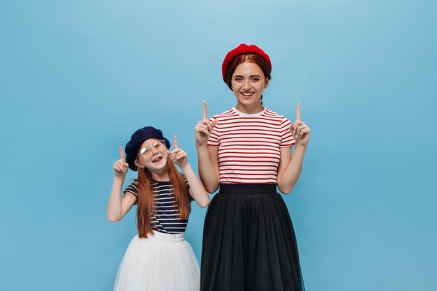 Zwei lustige Mädchen in coolen Baskenmützen und gestreiften schwarzen und roten T-Shirts lächeln und zeigen mit dem Daumen nach oben auf isoliertem Hintergrund