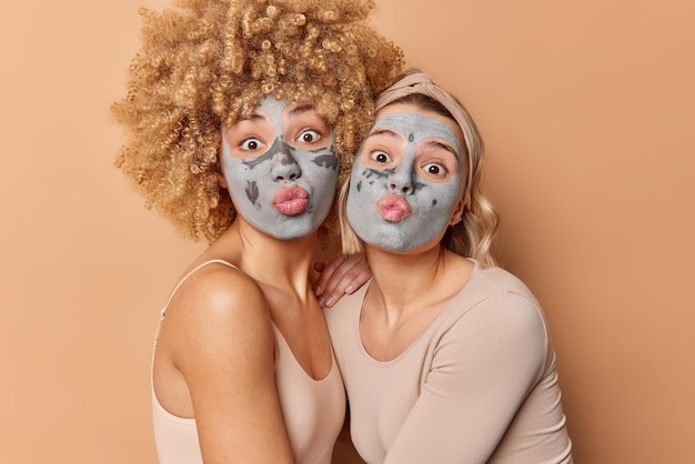 Zwei lustige weibliche Models halten die Lippen abgerundet, machen lustige Grimassen und tragen nährende Tonmasken auf das Gesicht auf, um sich zu verjüngen und zu verwöhnen, gekleidet lässig isoliert über braunem Hintergrund Beauty-Konzept