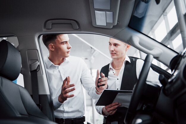 Zwei Männer stehen im Ausstellungsraum gegen Autos. Nahaufnahme eines Verkaufsleiters in einem Anzug, der ein Auto an einen Kunden verkauft. Der Verkäufer gibt dem Kunden den Schlüssel.