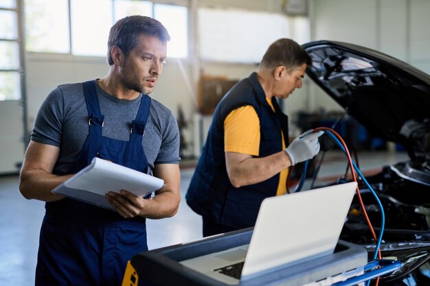 Zwei Mechaniker untersuchen die Fahrzeugpanne in der Autowerkstatt. Der Fokus liegt auf dem Mechaniker, der einen Laptop verwendet, während er Notizen in die Zwischenablage schreibt