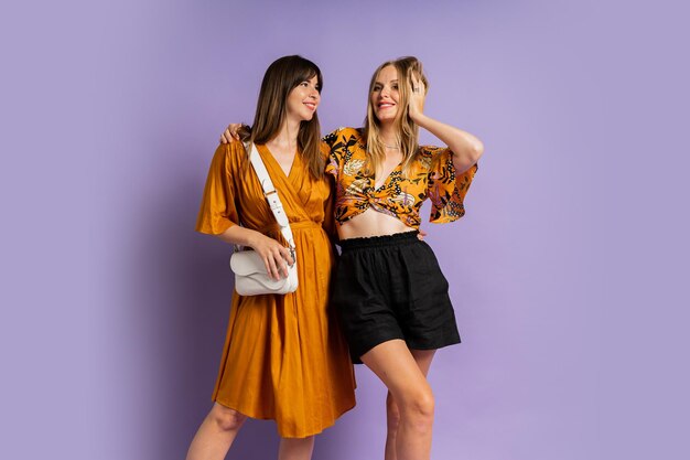 Zwei modische europäische Frauen posieren im Studio vor violettem Hintergrund und tragen ein stylisches Sommeroutfit