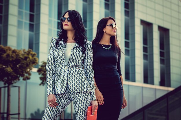 Zwei modische Frauen in stilvoller Kleidung und Sonnenbrillen posieren mitten im Geschäftsviertel.