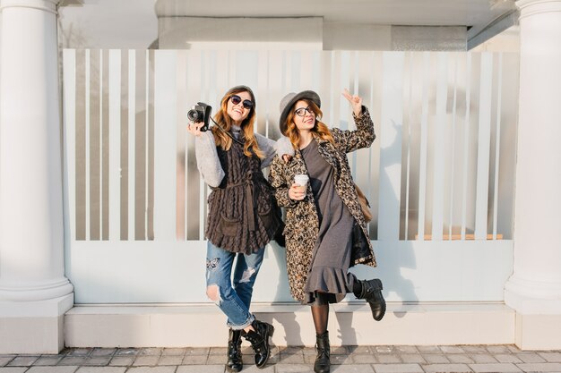 Zwei modische freudige lächelnde Frauen, die Spaß auf sonniger Straße in der Stadt haben. Stilvoller Look, zusammen reisen, moderne Trendkleidung tragen, mit Kaffee spazieren gehen und positive Emotionen ausdrücken.