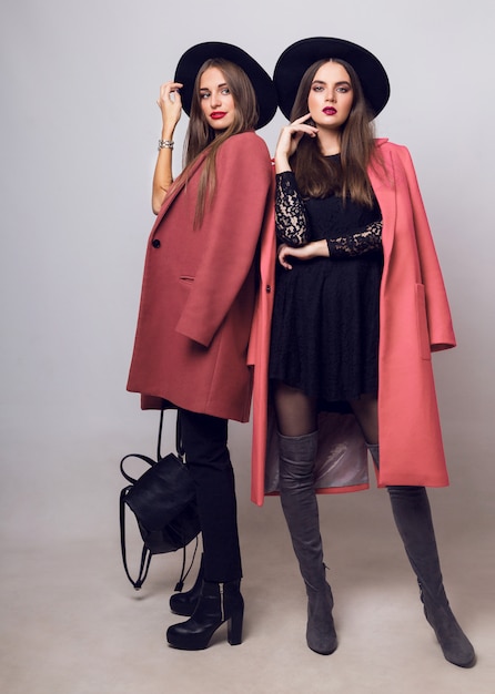 Zwei modische junge Frauen in lässigem, trendigem Frühlingsmantel, Stiefeln mit Absätzen, schwarzem Hut und stilvoller Handtasche
