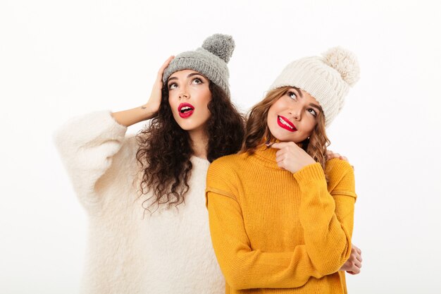 Zwei nachdenkliche Mädchen in den Strickjacken und in Hüten, die zusammen beim Schauen oben über weißer Wand aufwerfen