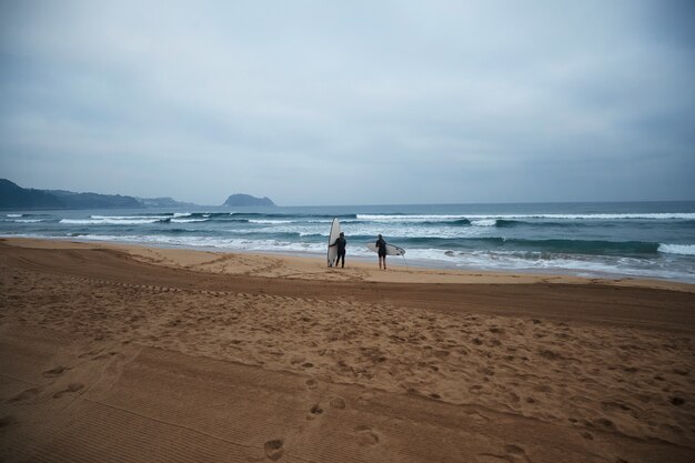 Zwei nicht wiederzuerkennende Surfmädchen mit ihren Longboards bleiben am Meer und beobachten am frühen Morgen Wellen, tragen volle Neoprenanzüge und sind bereit zum Surfen