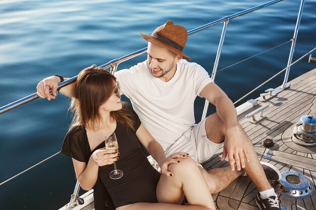 Zwei niedliche Leute in der Beziehung hängen auf der Yacht, sitzen auf dem Boden und sprechen, während sie mit Freunden zur Insel reisen. Das verliebte Paar reiste ins Ausland, um sich sorglos zu fühlen und sich zu amüsieren