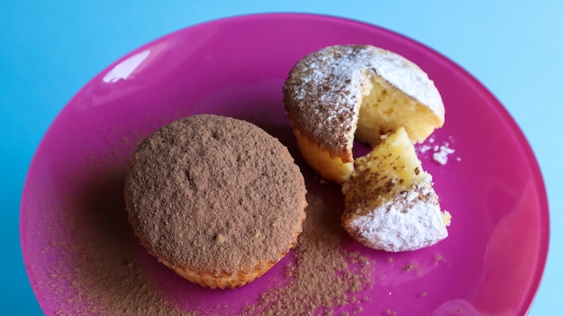 Zwei quarkkuchen bestreut mit schokolade und puderzucker auf einem rosa teller auf blauem grund. dessert, ein kleiner cupcake. weiße gebackene kekse mit einer luftigen textur. lebensmittelkonzept.