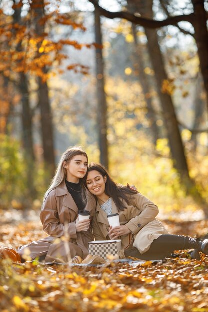 Zwei schöne Freundinnen verbringen Zeit auf einer Picknickdecke im Gras. Zwei junge lächelnde Schwestern machen ein Picknick und essen Croissants im Herbstpark. Brunette und blonde Mädchen, die Mäntel tragen.