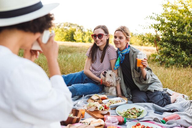 Zwei schöne Mädchen, die mit einem kleinen Hund auf einer Picknickdecke sitzen und vor der Kamera posieren, verbringen glücklich Zeit mit einem Picknick im Park