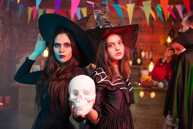 Zwei schöne mädchen in schwarzen kleidern und hexenhüten halten einen totenkopf für die halloween-party. fröhliche hexen.
