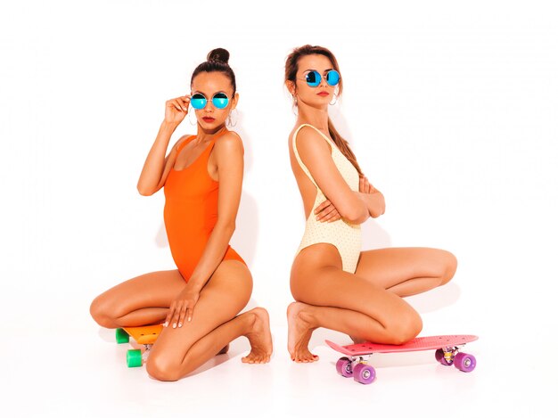 Zwei schöne sexy lächelnde Frauen in den bunten Badebekleidungsbadeanzügen des Sommers. Trendy Girls in Sonnenbrillen. Positive Modelle, die auf dem Boden mit bunten Penny-Skateboards sitzen. isoliert