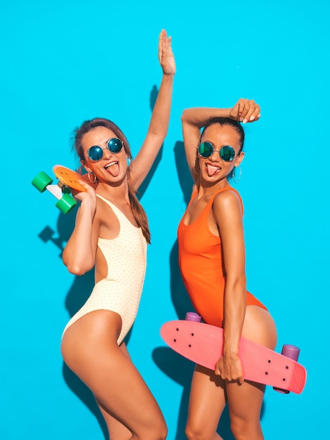Zwei schöne sexy lächelnde Frauen in den bunten Badebekleidungsbadeanzügen des Sommers. Trendy Girls in Sonnenbrillen. Positive Modelle, die Spaß mit bunten Penny-Skateboards haben. isoliert. Zeigt die Zunge