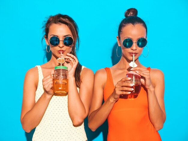 Zwei schöne sexy lächelnde Frauen in den bunten Badebekleidungsbadeanzügen des Sommers. Trendy Girls in Sonnenbrillen. Werde verrückt. Lustige Modelle isoliert. Frisches Cocktail smoozy Getränk trinken