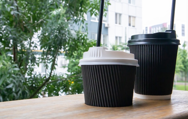 Zwei schwarze papierkaffeetassen mit deckeln im sommer im freien bei sonnigem wetter auf einem holztisch eines cafés, cafés oder restaurants.