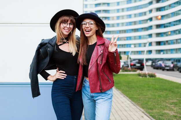 Zwei sorglos lächelnde Frauen, die auf der modernen Stadt aufwerfen. Tragen von Wollmütze, Lederjacke und Jeans.