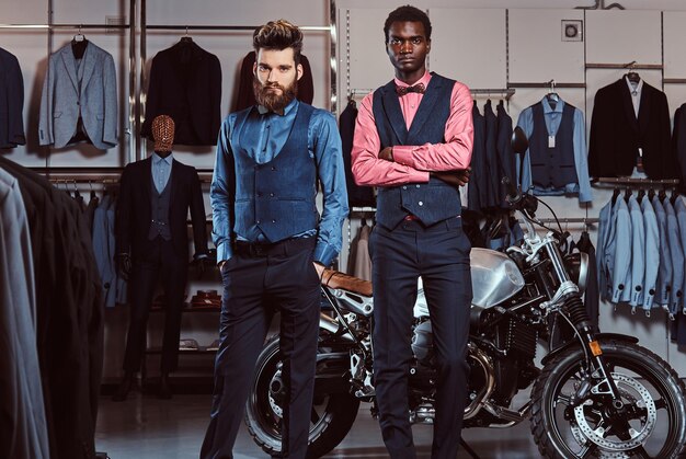 Zwei stilvolle, elegant gekleidete Männer posieren in der Nähe des Retro-Sportmotorrads im Herrenbekleidungsgeschäft.