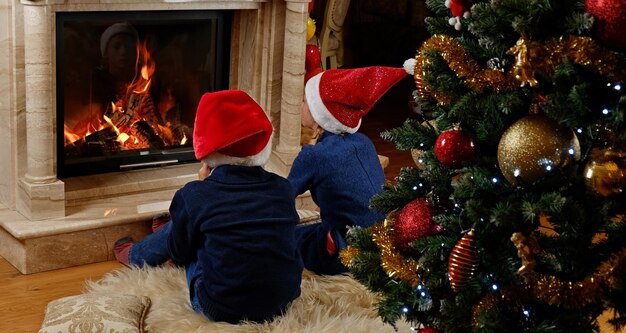 Zwei süße Kinder sitzen am Kamin im weihnachtlich dekorierten Raum.