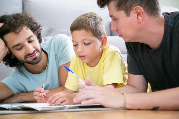 Zwei Väter helfen fokussierten Jungen bei der Hausaufgabe in der Schule, liegen zu Hause auf dem Boden, schreiben oder zeichnen in Papieren. Konzept der Familie und der schwulen Eltern