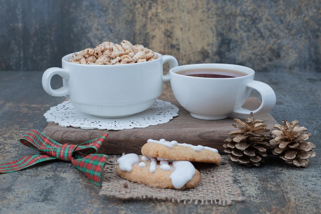 Zwei weiße tassen mit weihnachtsplätzchen und zwei tannenzapfen auf holzbrett.