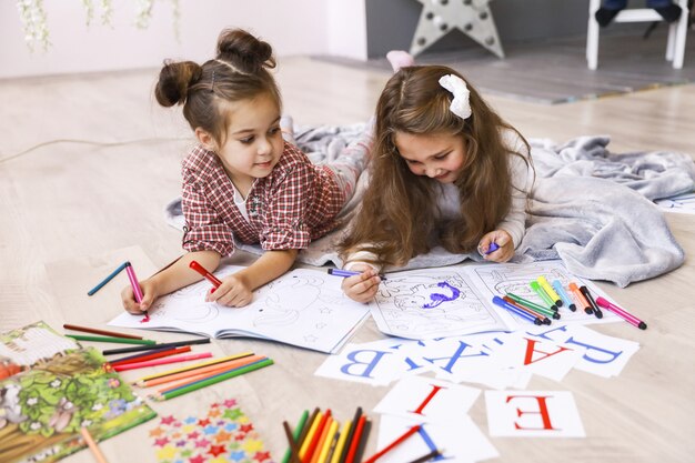 Zwei winzige glückliche Mädchen, die in dem Malbuch zeichnen, das auf dem Boden auf der Decke liegt und Buchstaben lernt
