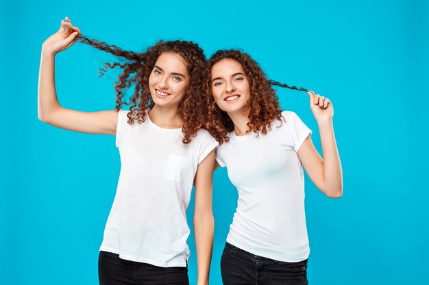 Zwei Zwillinge der Frau, die Haare halten und über Blau lächeln.