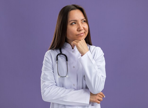 Zweifelhafte junge asiatische Ärztin, die medizinisches Gewand und Stethoskop trägt und die Hand am Kinn hält, isoliert auf lila Wand mit Kopierraum