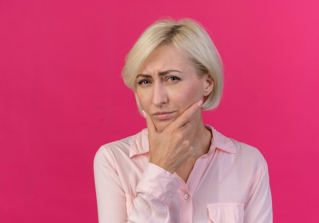 Zweifelhafte junge blonde slawische Frau, die Kamera und berührendes Kinn lokalisiert auf rosa Hintergrund mit Kopienraum betrachtet