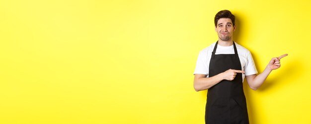Zweifelhafter Barista in schwarzer Schürze, der mit den Fingern nach rechts zeigt und skeptisch und unamüsiert übersteht