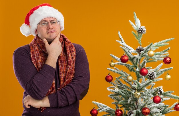 zweifelhafter erwachsener Mann, der Brille und Weihnachtsmütze mit Schal um den Hals trägt, der nahe verziertem Weihnachtsbaum steht und Hand auf Kinn hält, das Seite lokalisiert auf orange Hintergrund betrachtet