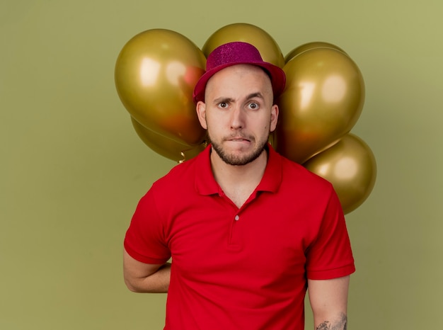 Zweifelhafter junger hübscher slawischer Party-Typ, der Partyhut hält, der Ballons hinter der beißenden Lippe des Rückens hält und Kamera lokalisiert auf olivgrünem Hintergrund mit Kopienraum betrachtet