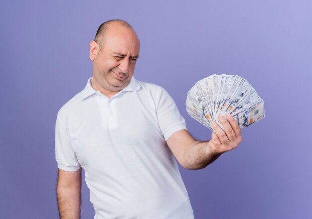zweifelhafter lässiger reifer Geschäftsmann, der Geld hält und betrachtet, das auf lila isoliert wird